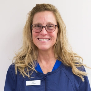 Dental professional wearing blue scrubs and glasses, smiling at camera in clinical setting.