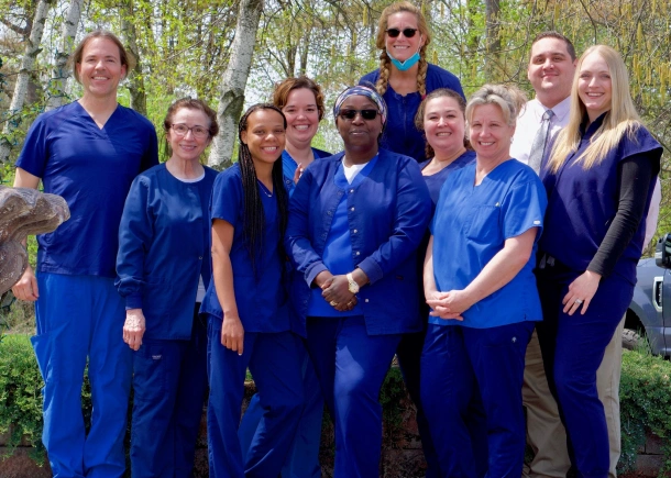 Dental team members in blue scrubs posing together outdoors among green trees.