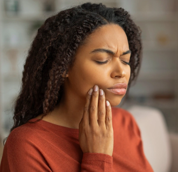 Woman in red shirt touching her cheek, appearing to experience dental pain or discomfort indoors.