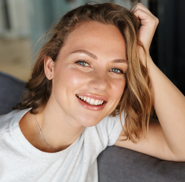 Young woman smiling at camera, wearing white shirt, hand in hair, indoor setting.
