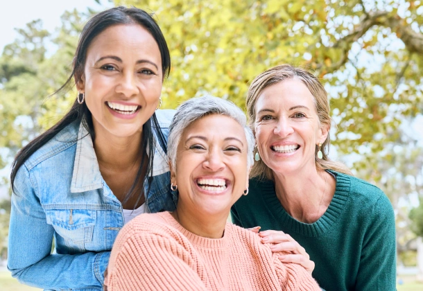 Three smiling women posing together outdoors with trees in background.