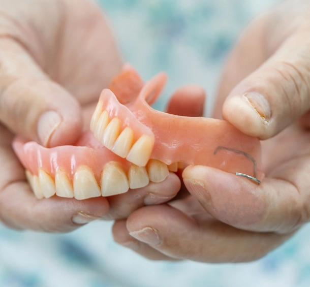 Hands holding a complete denture set displaying artificial teeth against a light blue background.