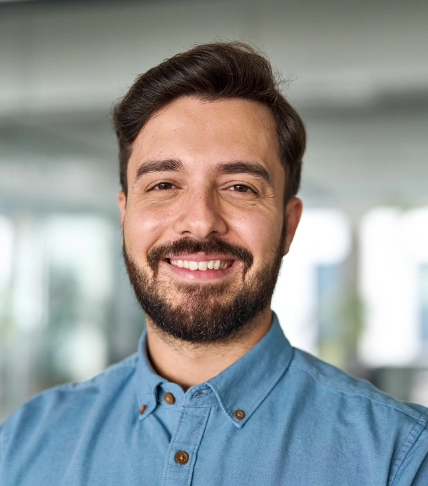 Professional headshot of smiling male dentist in blue shirt, modern dental office setting.