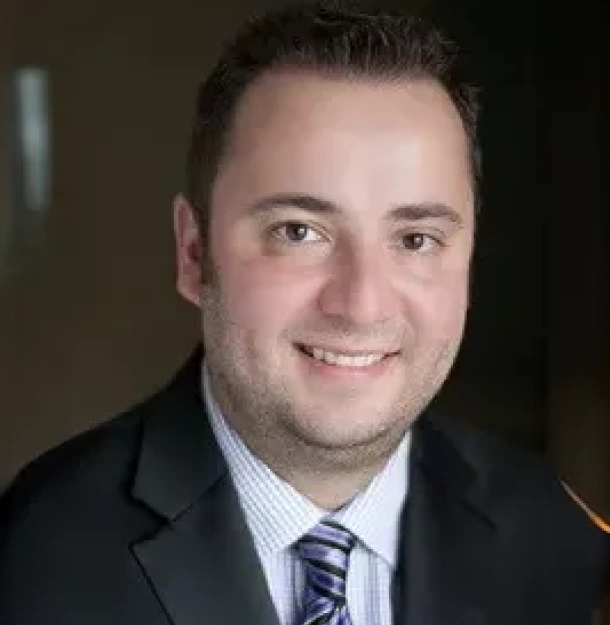 Professional headshot of a man in business attire smiling at camera, dental office setting.