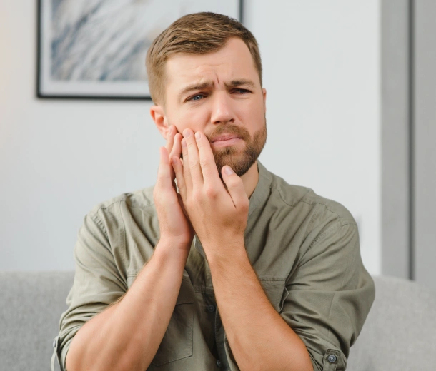Man holding jaw in pain, sitting indoors in dental office setting.