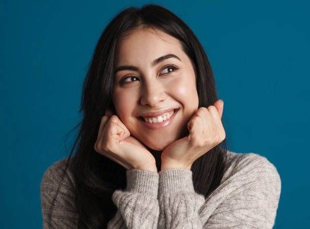 Smiling woman with dark hair resting chin on hands against blue background, dental practice portrait.