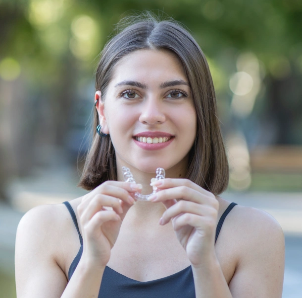 Young woman smiling outdoors holding clear dental aligner in front of blurred green background.
