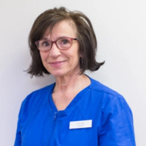 Female dental professional wearing blue scrubs and glasses, smiling at camera in clinical setting.