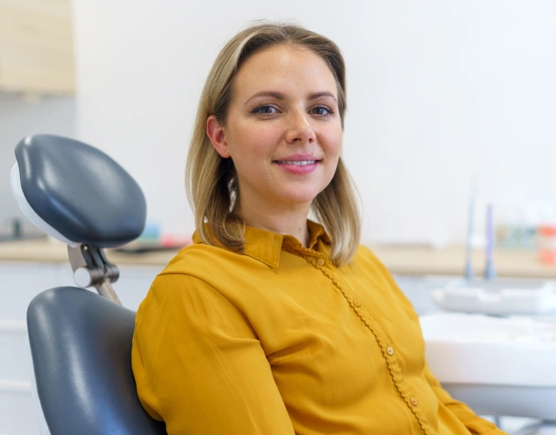 Woman in yellow shirt smiling at dental office with chair visible in background.
