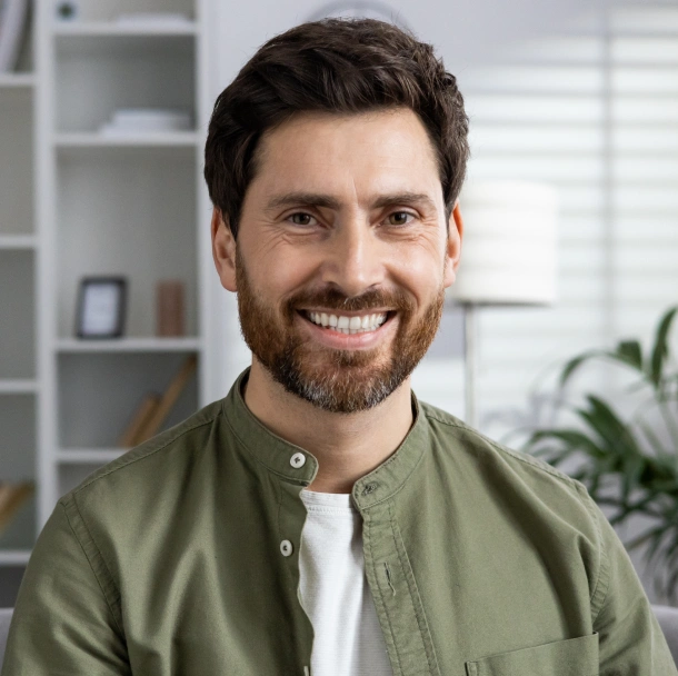 Smiling male dental professional in sage green shirt, modern office setting with shelving background.
