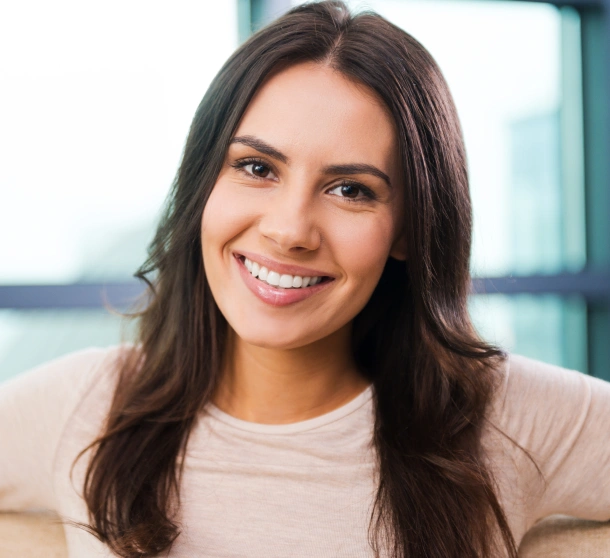 Woman smiling at camera in bright office setting, wearing light beige shirt.