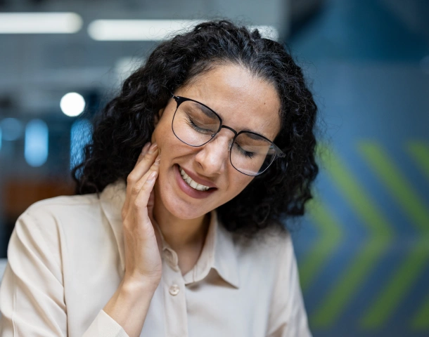Smiling woman wearing glasses in professional dental office setting, touching her face.