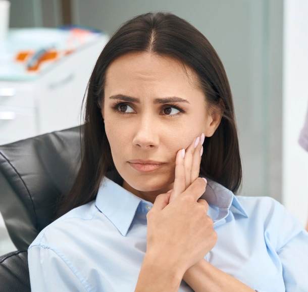 Woman in light blue shirt sitting at desk in office, hand to face, appearing thoughtful or concerned.