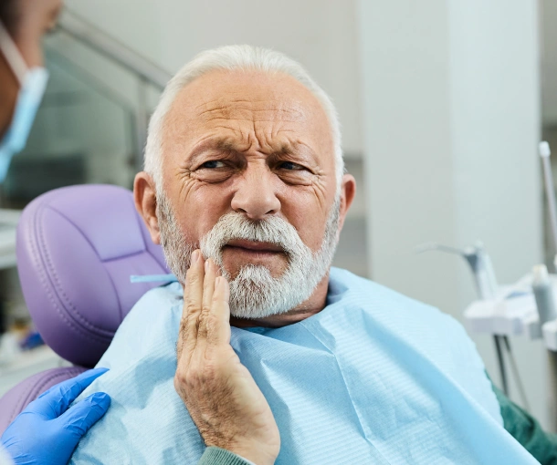 Senior man with toothache touching face in dental office chair wearing bib