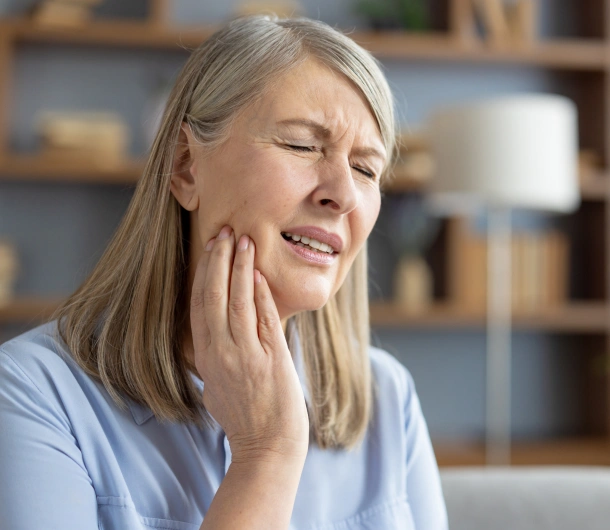 Middle-aged woman touching her cheek in pain, sitting indoors with shelving in background.